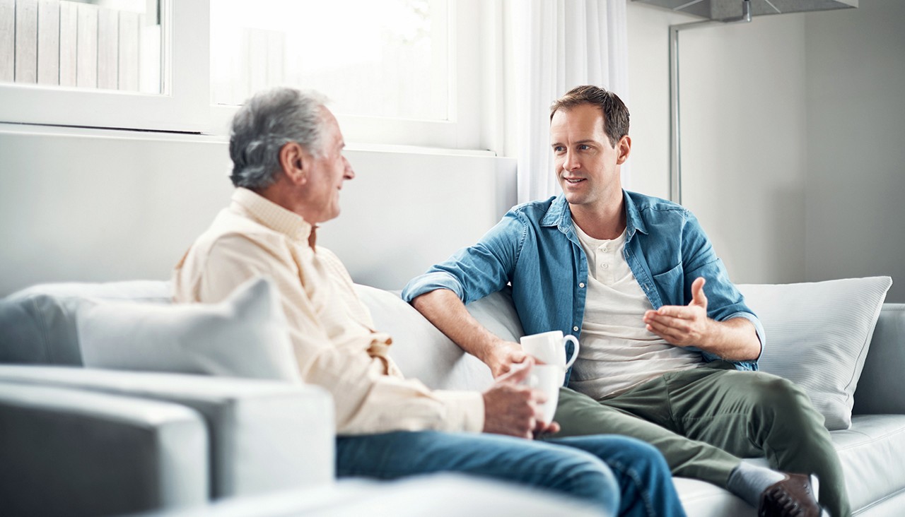 A younger man sitting on a couch talking to an older man as they drink coffee.