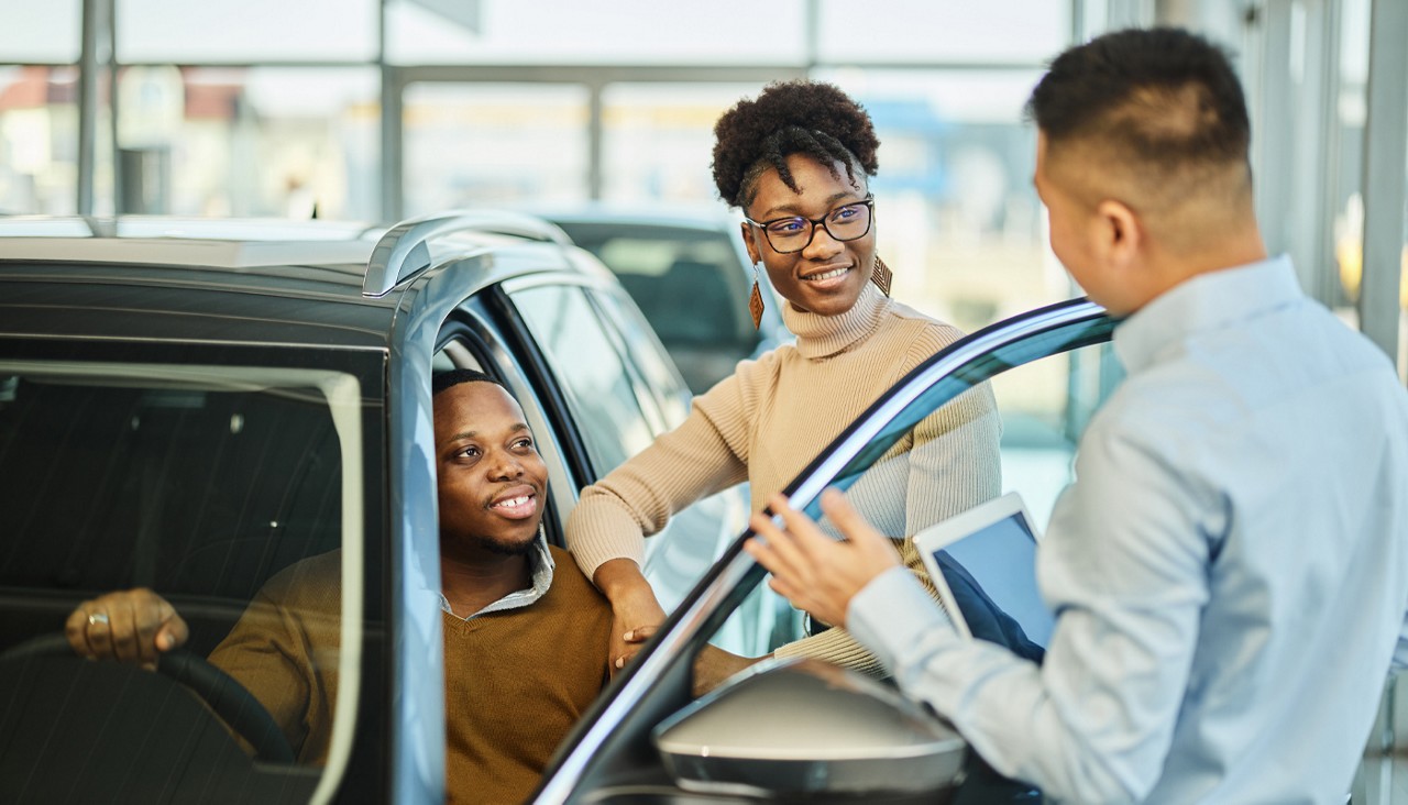 Young couple talking to a car salesman as they sit and stand in an open door to a new car.