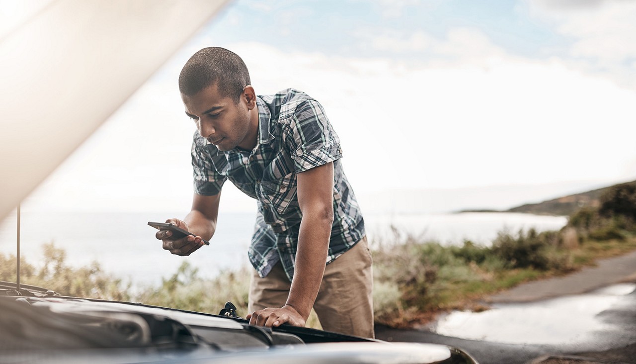 Person looking under hood of car on side of road, holding smartphone