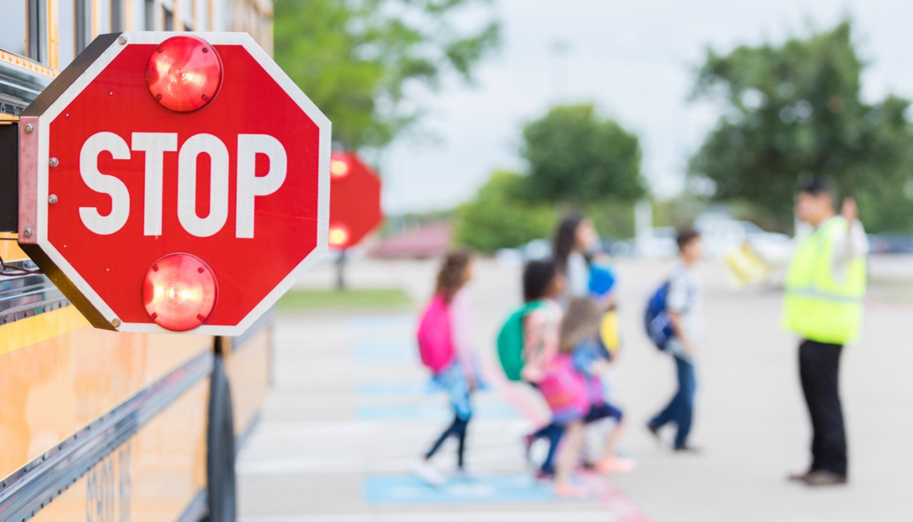 A close up of a school bus stop sign with children crossing the street in the background.