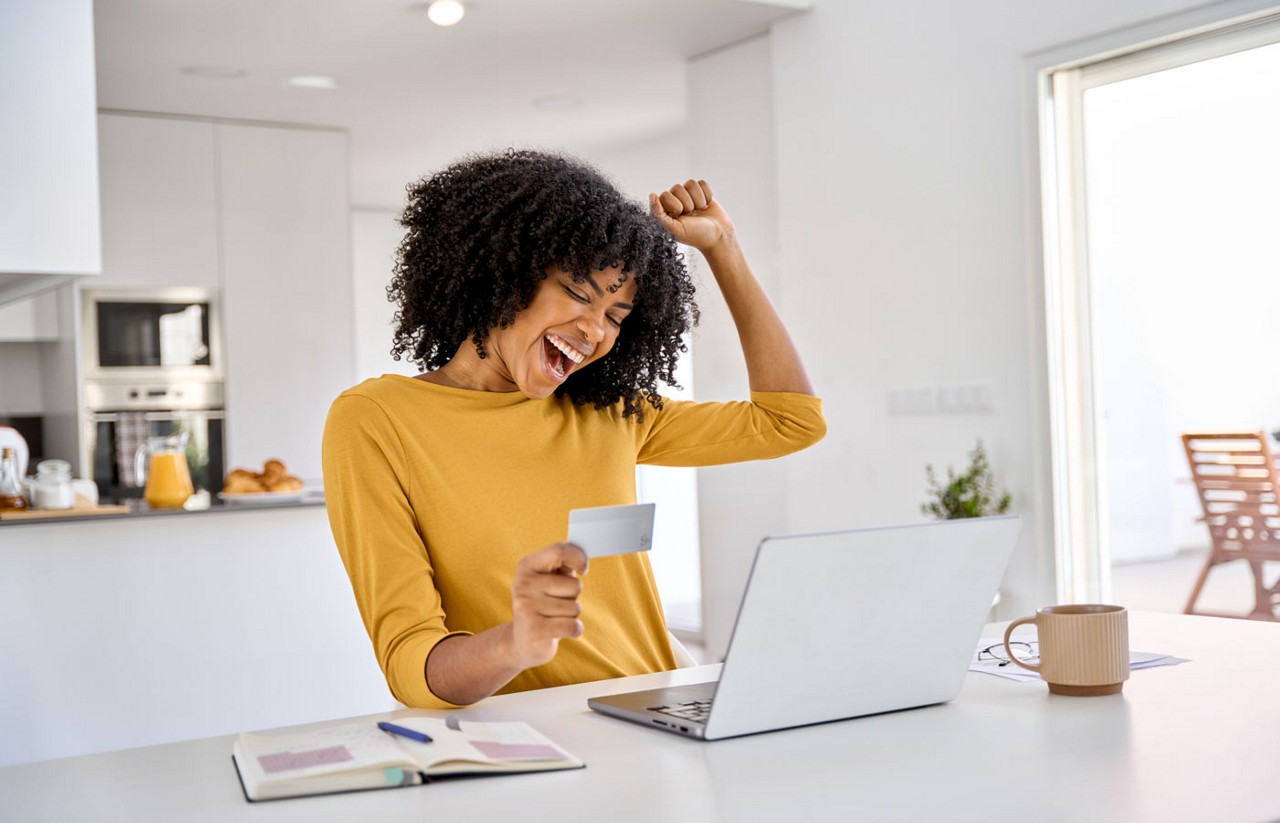 Happy excited young African woman customer winner holding credit card using laptop computer in kitchen celebrating money win receiving cashback getting online bonus for shopping sitting at home.