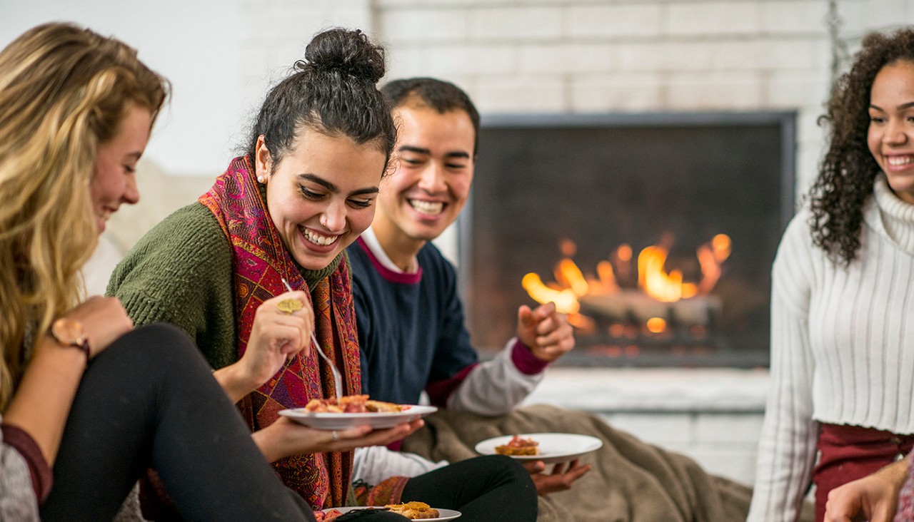 A group of friends eating near a fireplace with a roaring fire.