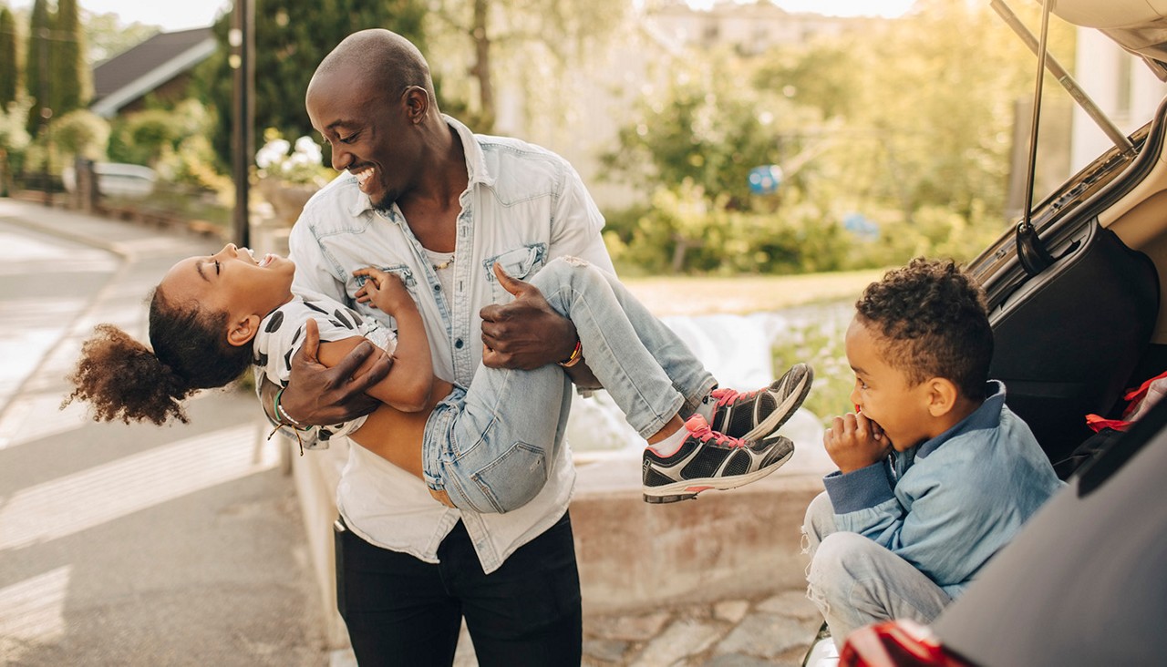 A father holding a laughing girl while a young boy watches from the car.