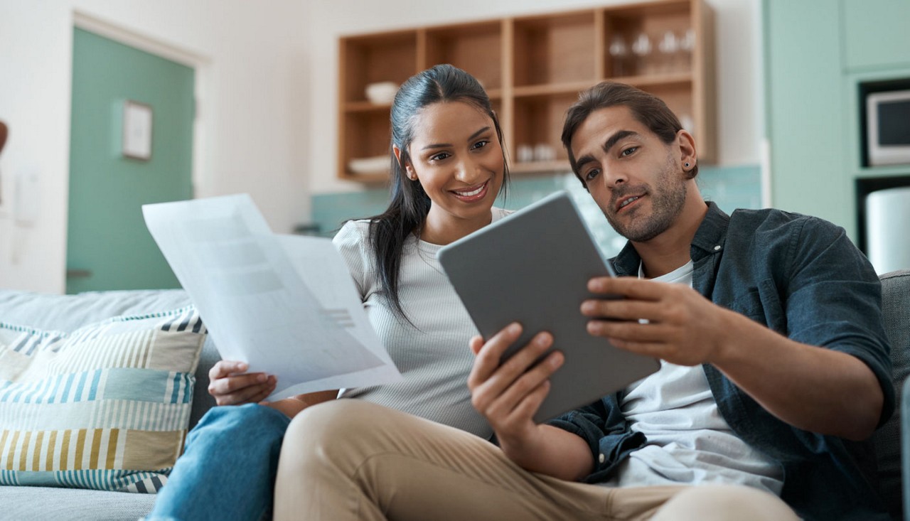 A young couple sitting on their couch looking at a tablet and paper together.