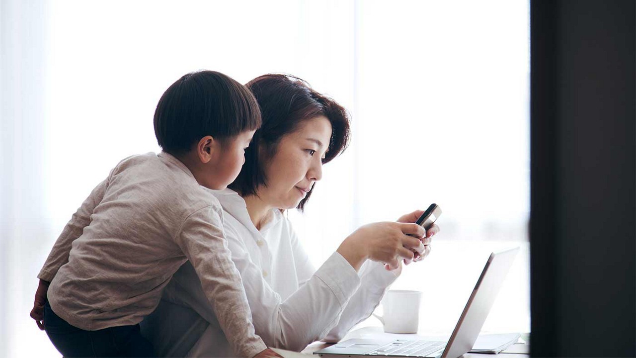 A mother and her son looking at a computer screen and phone.