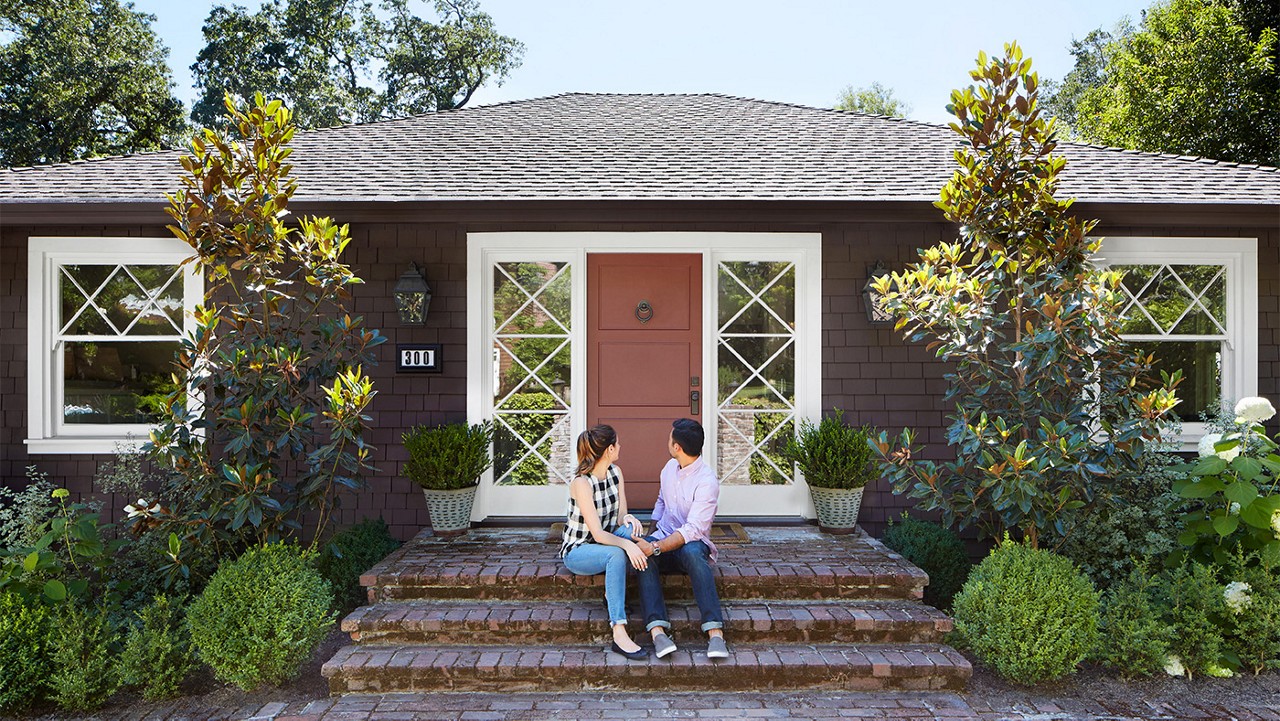 A young couple sitting on the front steps of their small brown cottage home.