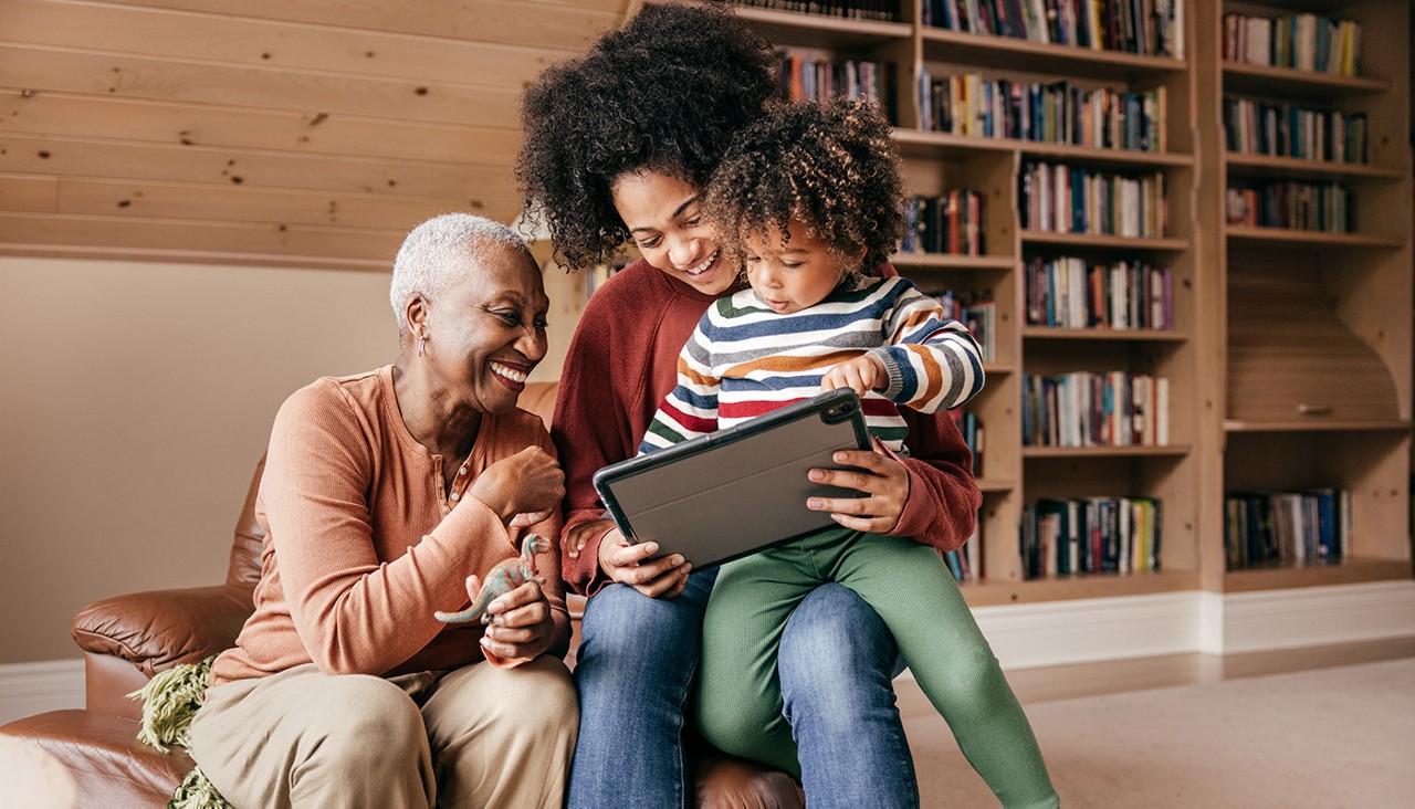 Grandmother, mother and child sit together smiling, looking at a tablet