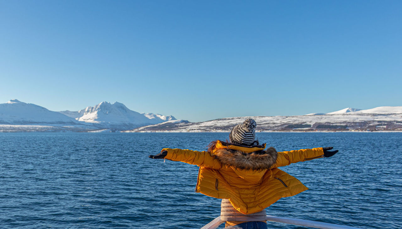 A woman wearing a yellow jacket with her arms out stretched admiring the Alaskan coastline.