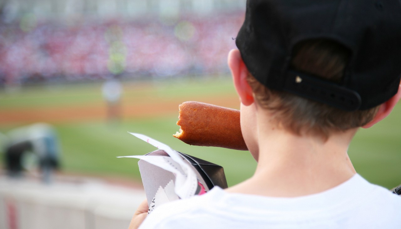 A young boy watching a baseball game while eating a corn dog.