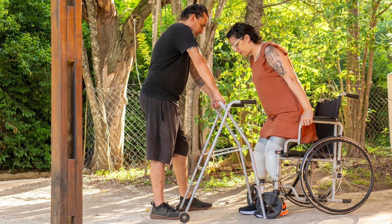 Man assisting woman with prosthetic legs using walker in rehabilitation center garden