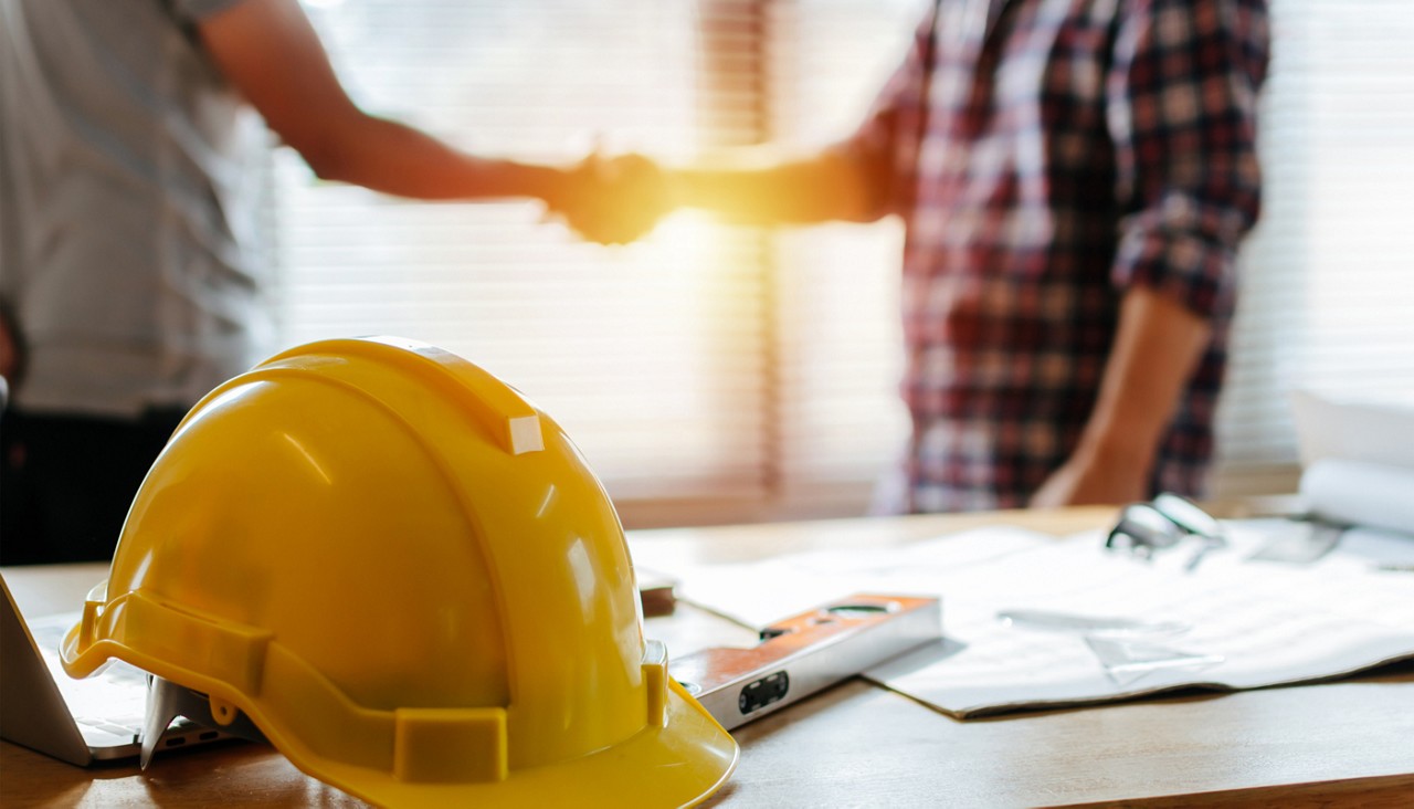 Yellow safety helmet on table and construction worker shaking hands with homeowner in background 