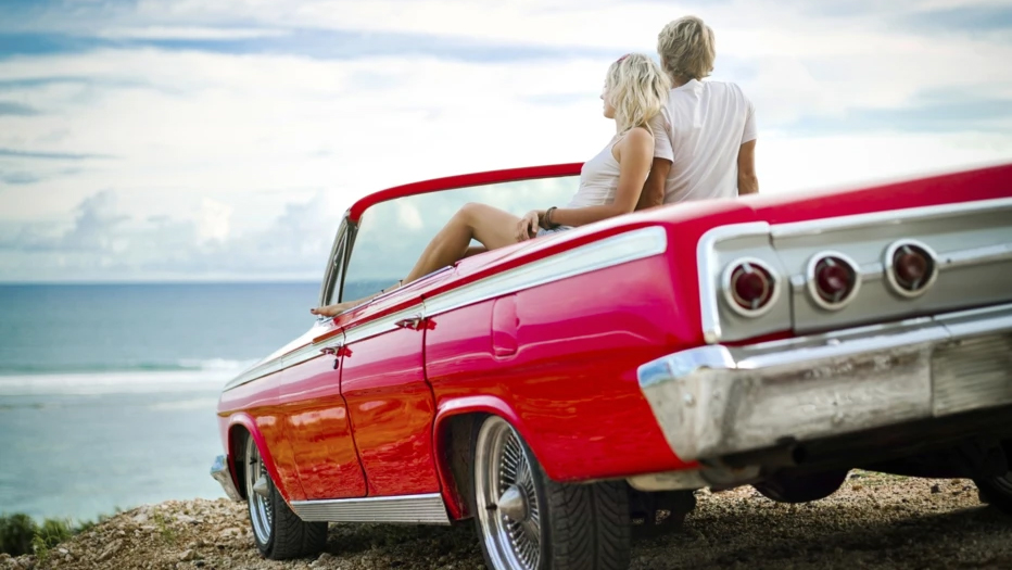 A man and a woman sitting in a parked red convertible on a cliff overlooking an ocean.