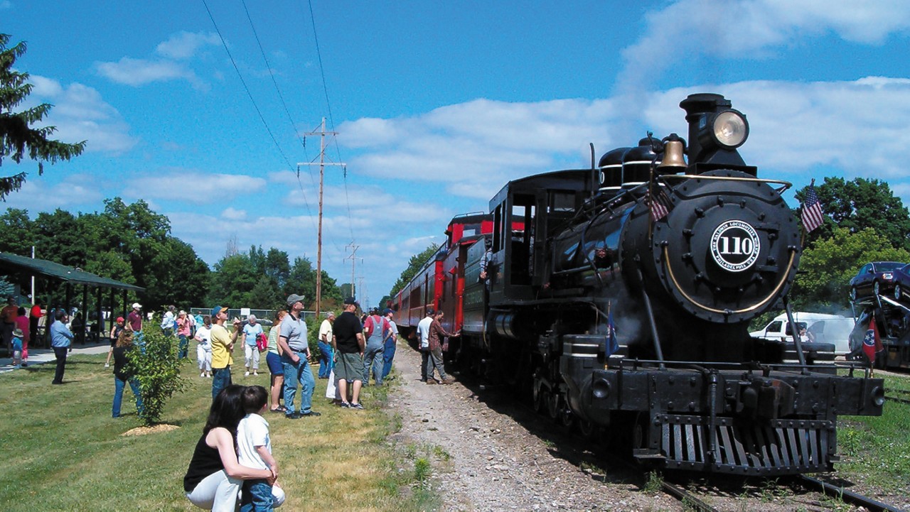 A group of people standing next to railroad tracks and the little River Railroad, a vintage steam locomotive.