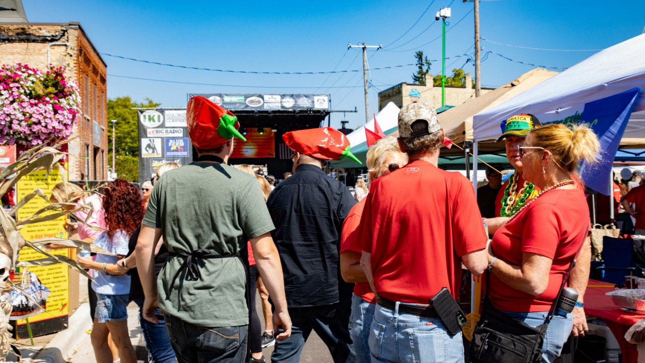 Chilimania festival scene with people in red shirts and chili pepper hats. Tents line the street, and a music stage is visible under a clear blue sky. 