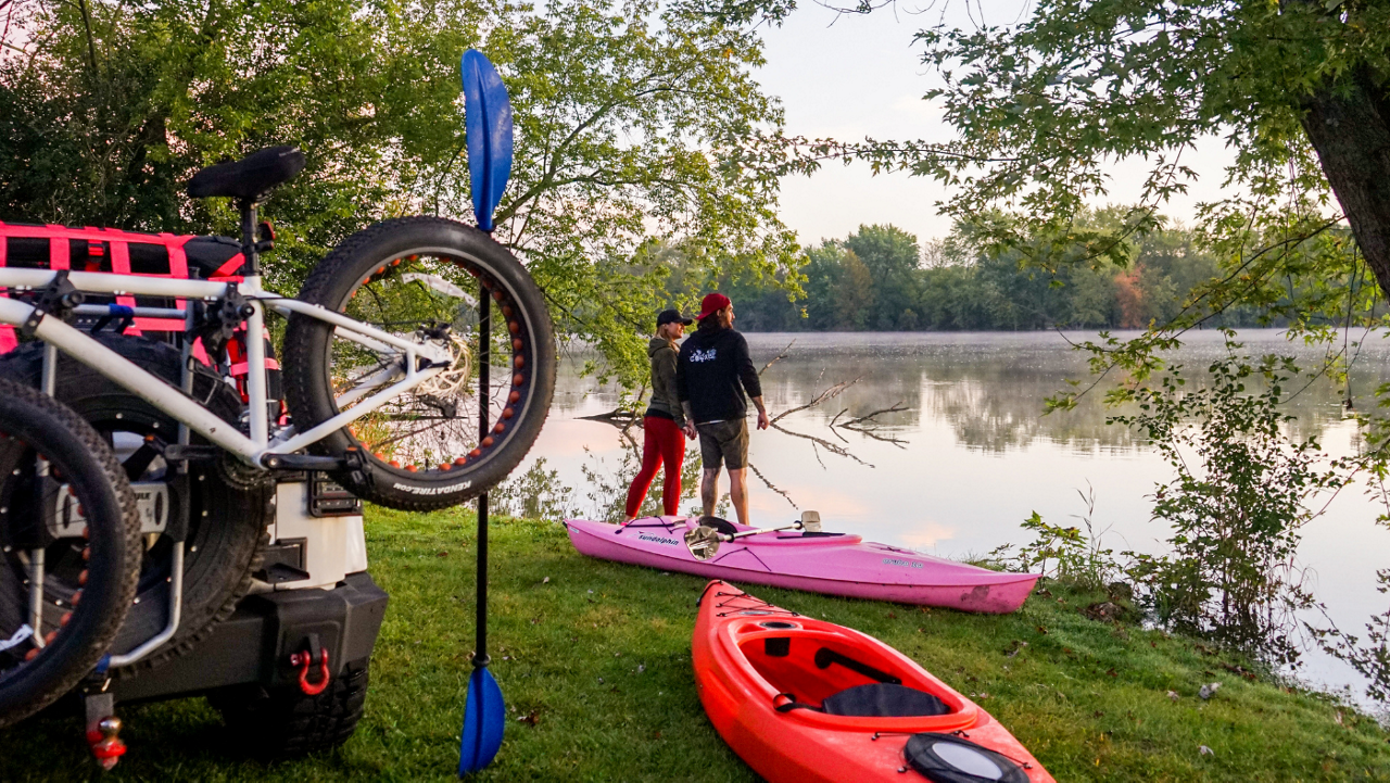 A couple in kayaking gear standing on a lakefront with a pink kayak and a red kayak with a vehicle in the background with bikes attached to the back.