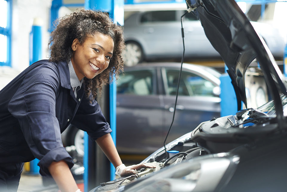 A female mechanic is working under a bonnet of a car in a garage repair shop. She is wearing blue overalls.  She is looking proudly  to camera.
